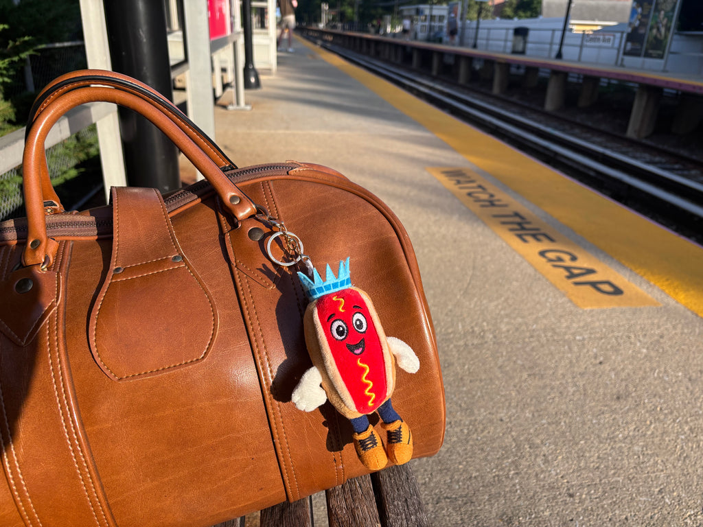 Empire Frank plush charm clipped to a leather bag on a train platform